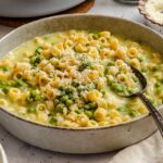 Wide view of a bowl of pasta e piselli topped with grated Parmesan, served with a spoon and extra cheese on the side.