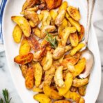 Roasted fingerling potatoes tossed with rosemary and garlic, served on a white platter with a spoon and coarse salt on the side.