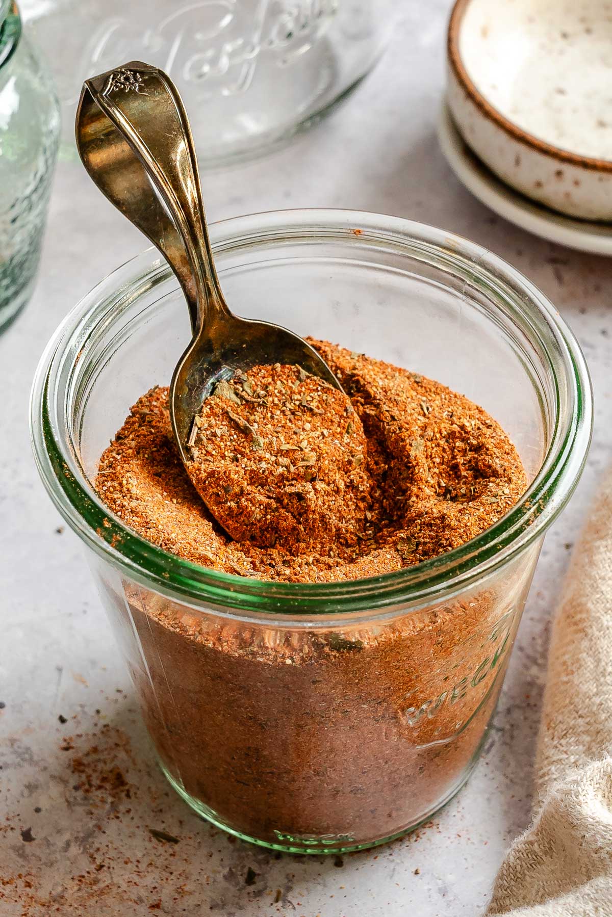 Angled view of a glass Weck jar generously filled with homemade Cajun creole seasoning, a vintage spoon dipped into the reddish-brown spice blend, with a linen cloth and empty jar nearby.
