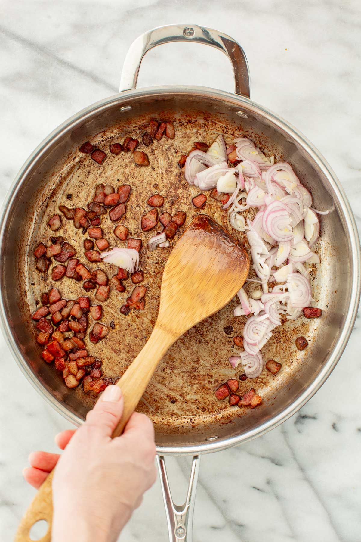 Crisped pancetta pushed to one side of a stainless steel skillet, with sliced shallots added to the other side, being stirred with a wooden spoon.