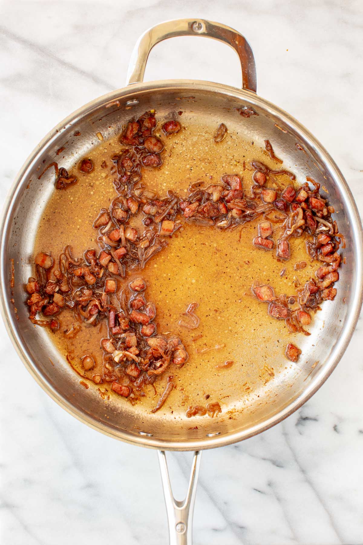 Caramelized shallots and pancetta in a golden-brown pan sauce in a stainless steel skillet.