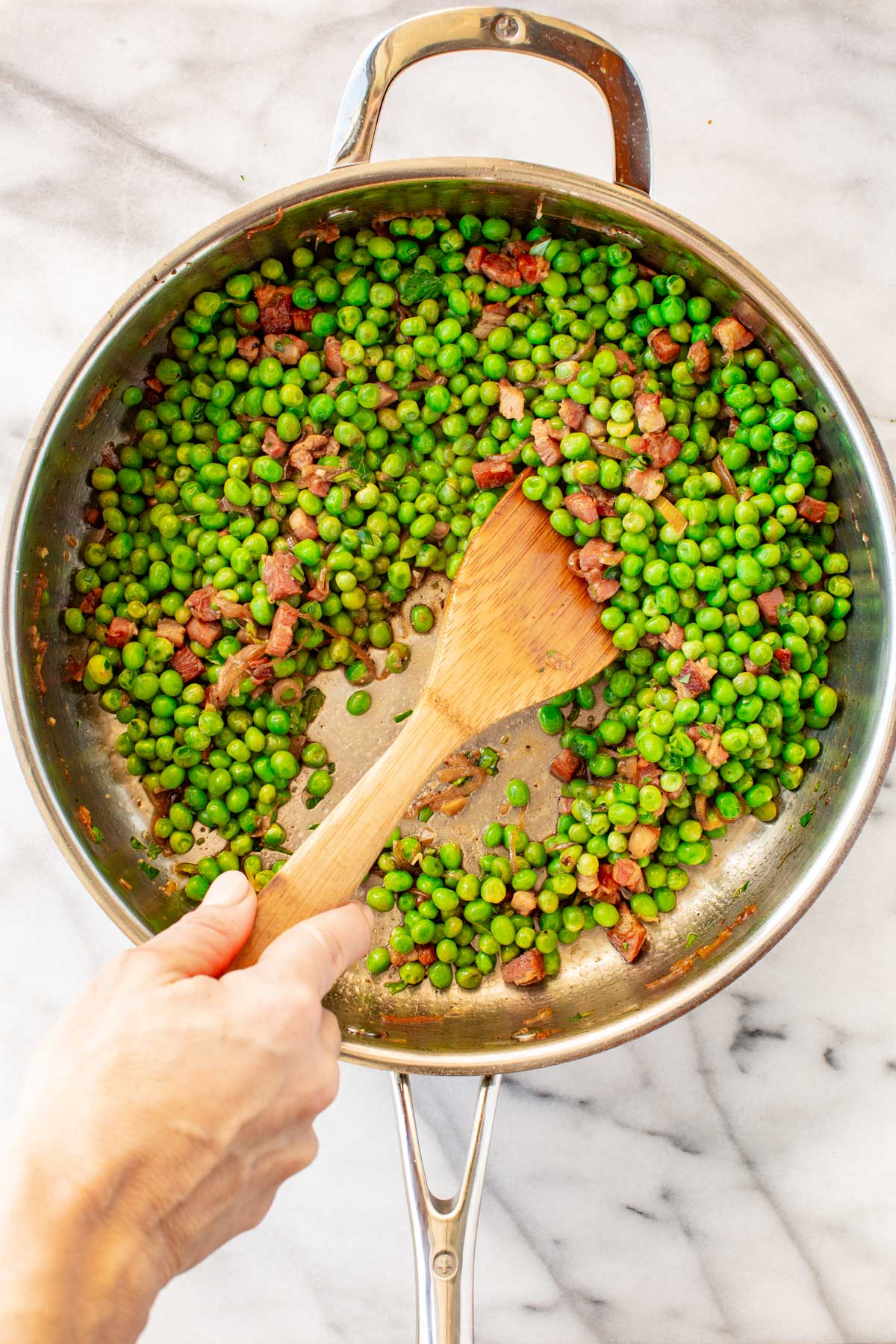Bright green peas being stirred together with crispy pancetta and caramelized shallots using a wooden spoon in a skillet.