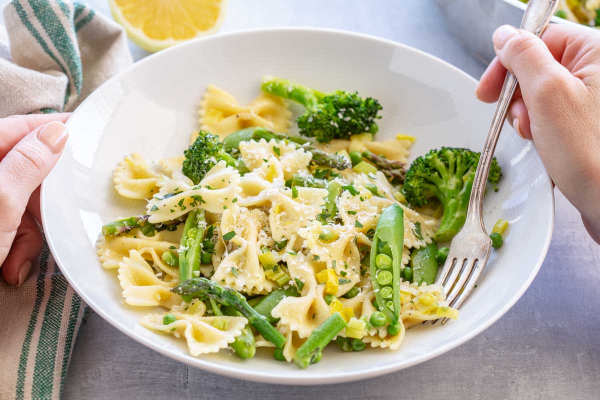 A pair of hands holding a white bowl of pasta primavera with a fork, showing farfalle pasta mixed with broccoli, asparagus, snap peas, peas, and leeks, dusted with parmesan and fresh herbs, with a lemon half and striped napkin in the background.