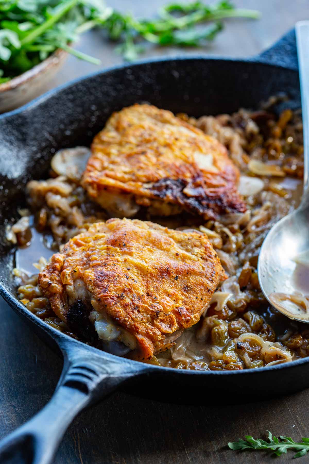 Close-up shot of two seared chicken thighs with deeply golden, crisped skin nestled in a rich, jammy agrodolce sauce of caramelized shallots and golden raisins in a black cast iron skillet, with a serving spoon resting alongside and arugula visible in the background.