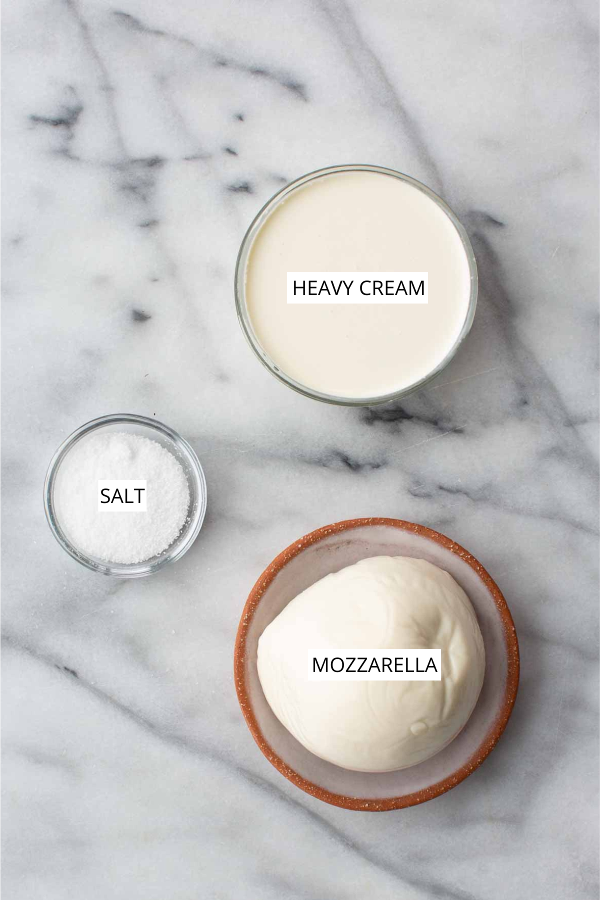 Overhead shot of stracciatella ingredients on a marble surface: a glass bowl of heavy cream, a small glass bowl of salt, and a ball of fresh mozzarella in a terracotta dish, each labeled.