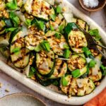 Overhead view of a white oval platter of grilled zucchini with shaved Parmigiano, toasted pine nuts, and fresh basil, with silver serving tongs, a bowl of pine nuts, a wedge of Parmigiano, and an orange napkin arranged around it.