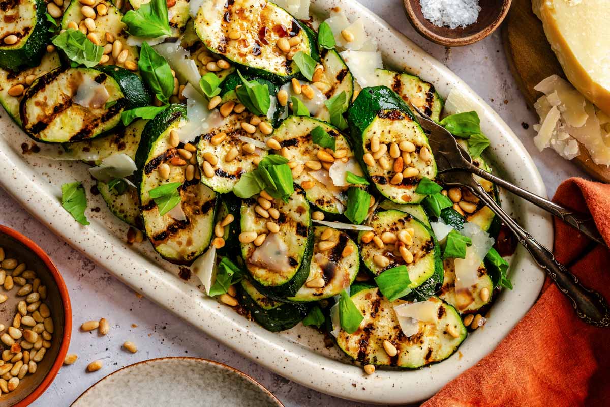 Overhead view of a white oval platter of grilled zucchini with shaved Parmigiano, toasted pine nuts, and fresh basil, with silver serving tongs, a bowl of pine nuts, a wedge of Parmigiano, and an orange napkin arranged around it.