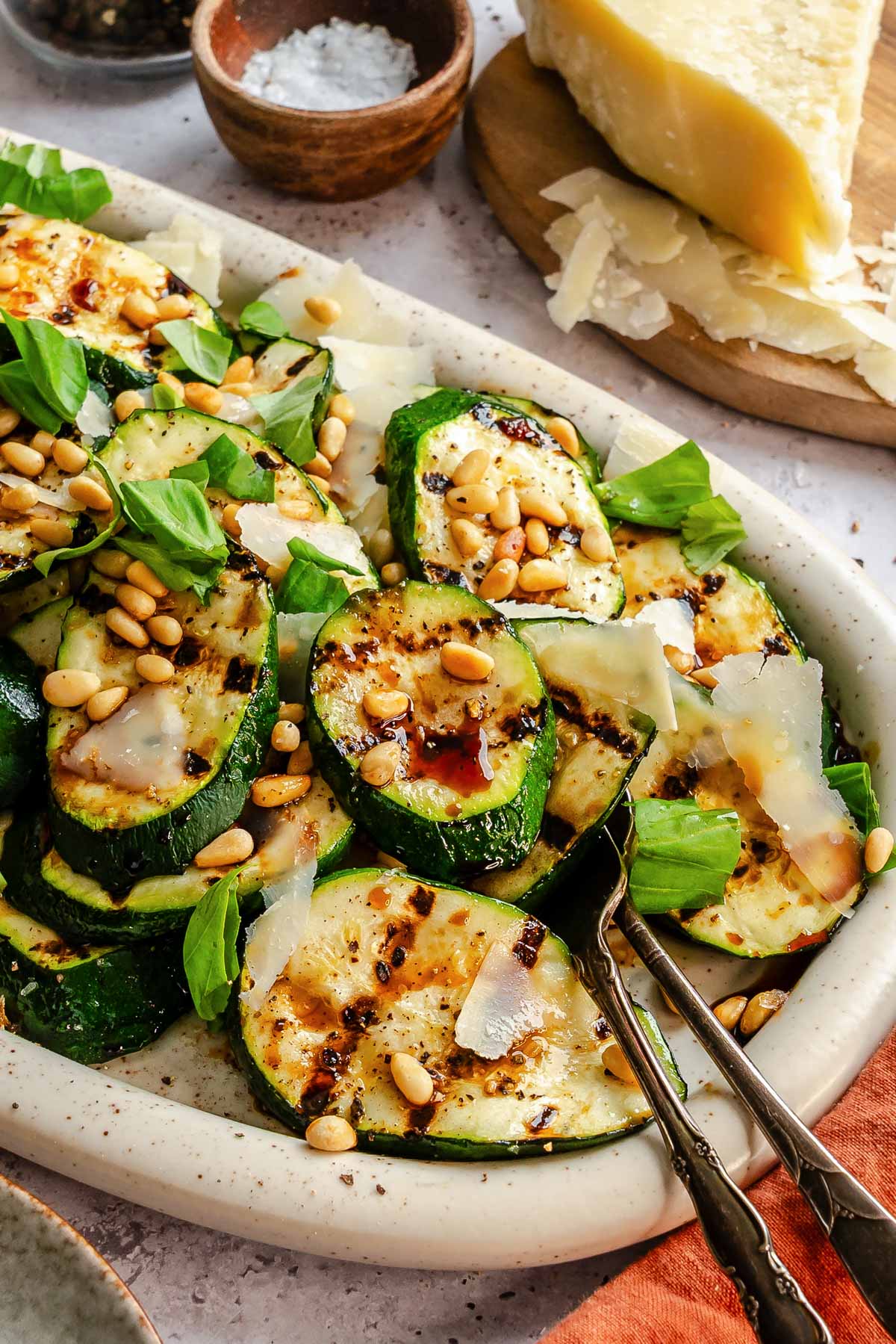 Side-angle view of a white speckled platter of grilled zucchini rounds topped with shaved Parmigiano, toasted pine nuts, fresh basil, and balsamic drizzle, with a wedge of Parmigiano and small wooden bowls of flaky salt and black pepper in the background.