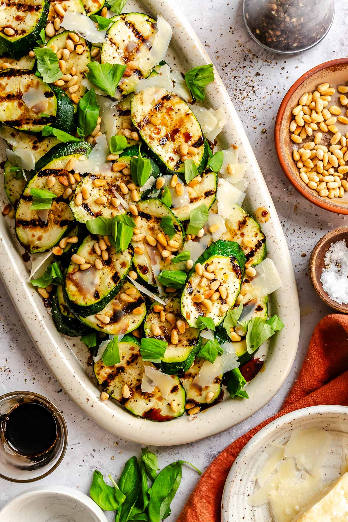 Overhead view of a white speckled oval platter with grilled zucchini rounds layered with shaved Parmigiano, toasted pine nuts, and torn fresh basil, with serving tongs resting on the side and small bowls of ingredients arranged around the platter.