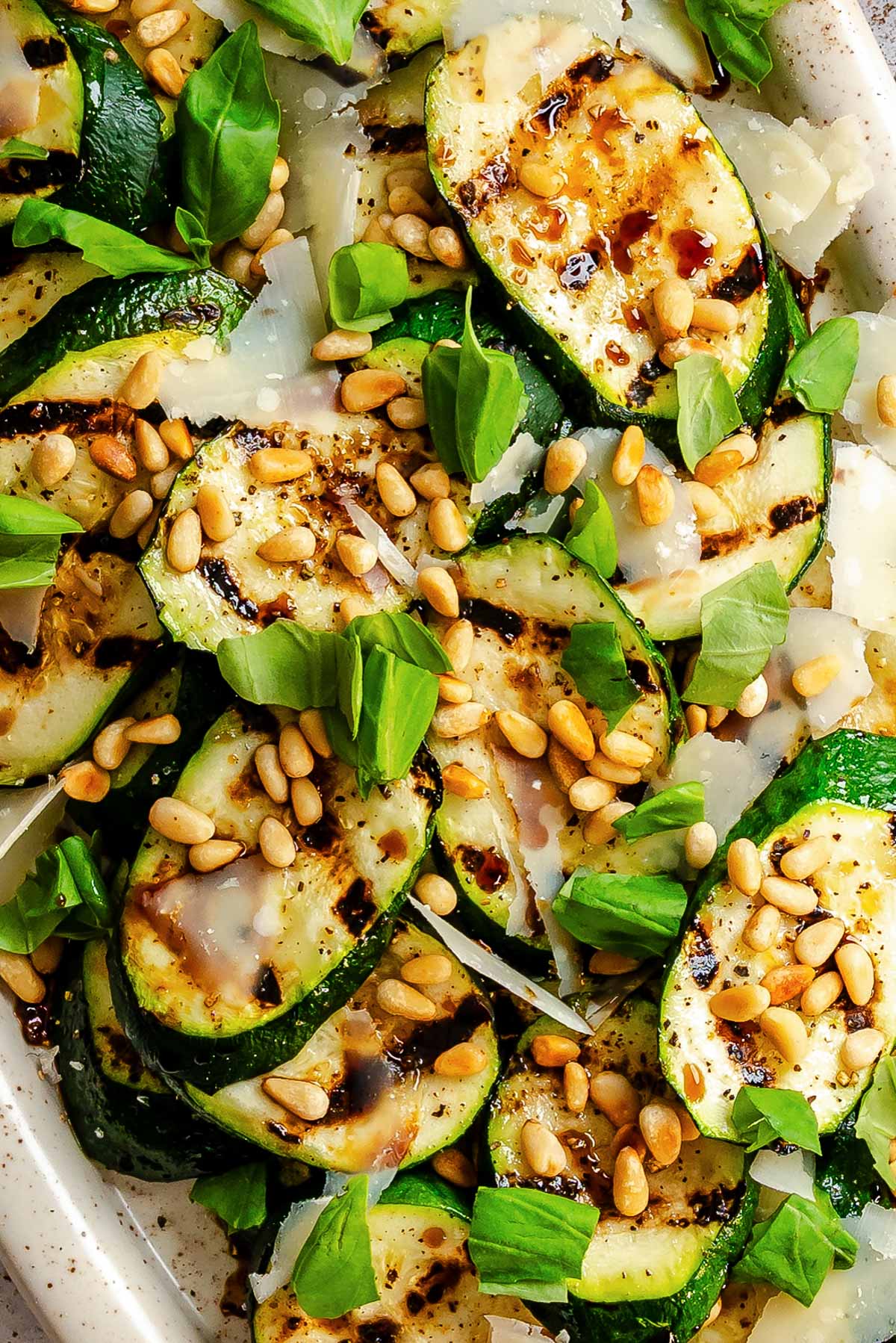Close-up of grilled zucchini rounds piled on a white platter, showing charred grill marks and topped with shaved Parmigiano, toasted pine nuts, fresh basil leaves, and drops of aged balsamic vinegar.
