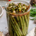 A filled Weck-style glass jar of pickled green beans, sealed and resting on a light stone surface surrounded by scattered props. The beans are packed vertically and the brine has turned slightly amber. The top of the jar is crowded with softened bean pieces, coriander seeds, garlic cloves, and wilted dill. Surrounding the jar: a linen napkin, a silver fork, scattered peppercorns, a halved garlic head, fresh dill sprigs, a jar of mixed peppercorns, and a Weck lid with orange rubber seal.