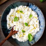 Overhead shot of a bowl of stracciatella cheese on a rustic wooden table, drizzled with olive oil, topped with basil leaves and black pepper, with a wooden spoon, bread slices, and cherry tomatoes nearby.