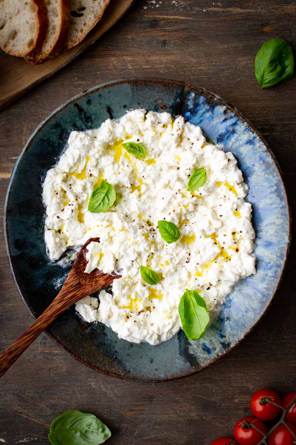 Overhead shot of a bowl of stracciatella cheese on a rustic wooden table, drizzled with olive oil, topped with basil leaves and black pepper, with a wooden spoon, bread slices, and cherry tomatoes nearby.