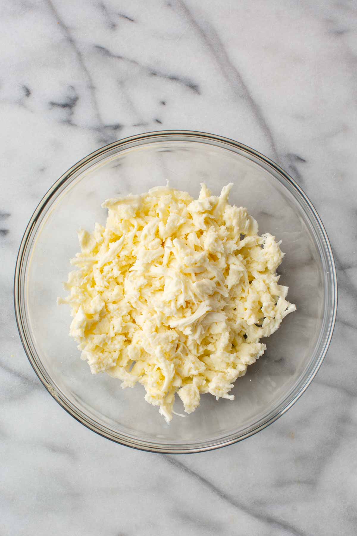 Overhead view of a glass bowl filled with shredded mozzarella curds, ready to be mixed with cream.