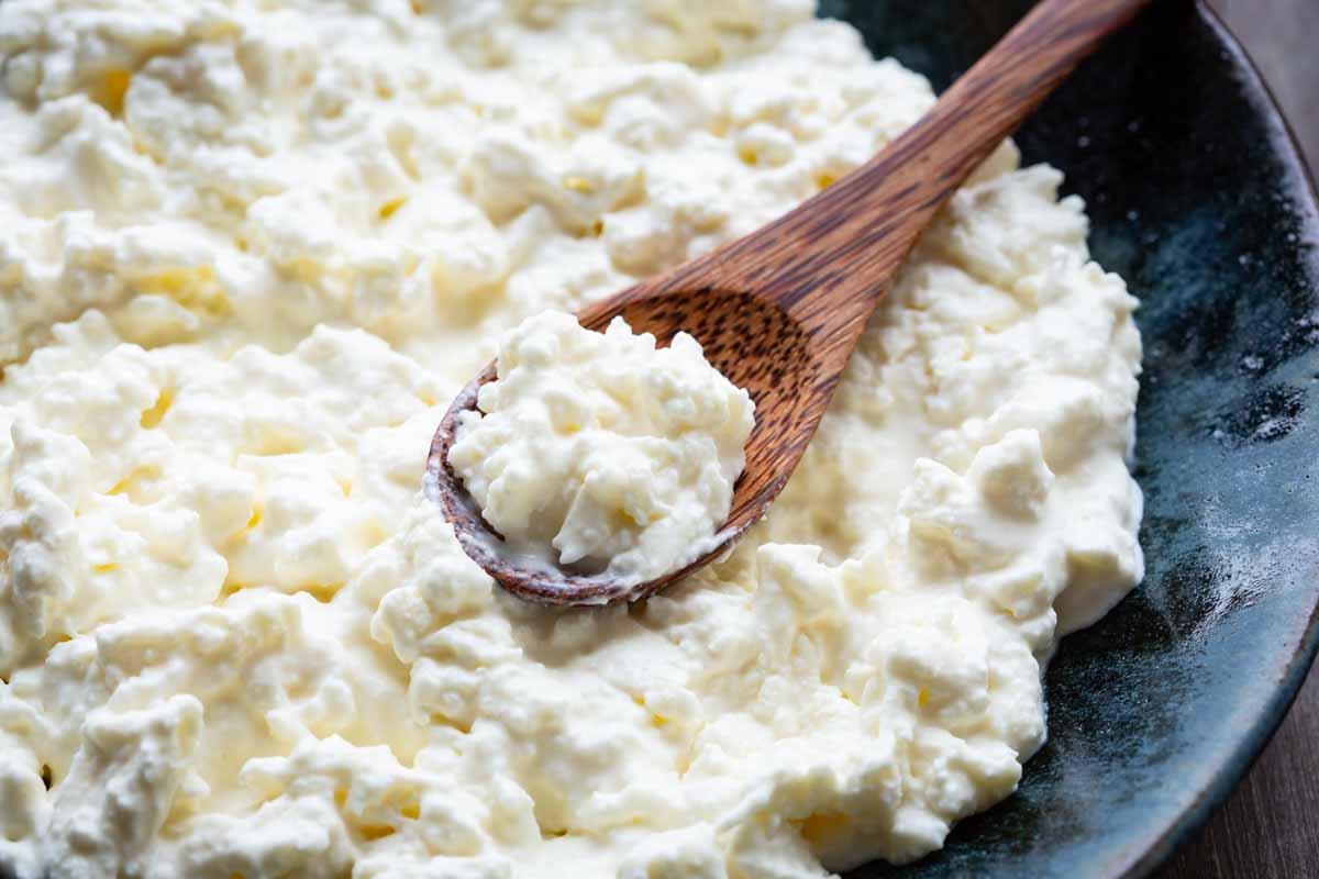 Close-up of finished stracciatella cheese piled on a dark plate, with a wooden spoon scooping up a portion of the creamy curds.