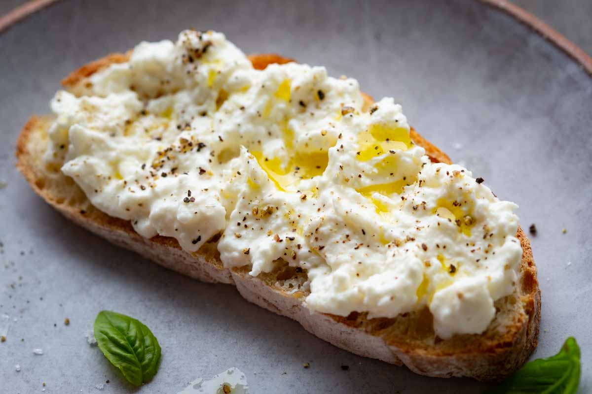 Close-up of a toasted bread slice generously spread with creamy stracciatella, drizzled with olive oil and topped with cracked black pepper and fresh basil.