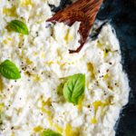 Close-up overhead view of stracciatella cheese in a dark bowl, drizzled with olive oil, sprinkled with black pepper, and garnished with fresh basil leaves, with a wooden spoon resting in it.
