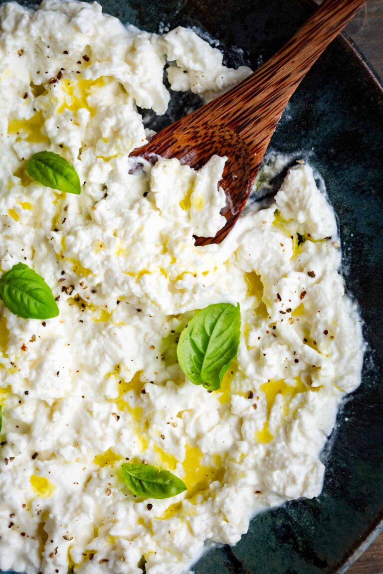 Close-up overhead view of stracciatella cheese in a dark bowl, drizzled with olive oil, sprinkled with black pepper, and garnished with fresh basil leaves, with a wooden spoon resting in it.