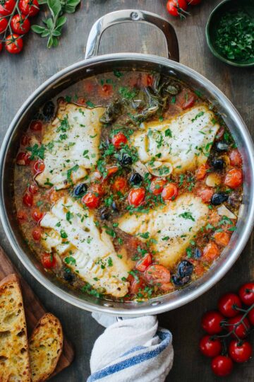 Overhead shot of fish acqua pazza in a stainless steel skillet, with four white fish fillets simmered in a tomato and olive broth, garnished with fresh parsley, surrounded by cherry tomatoes, oregano, and toasted bread.