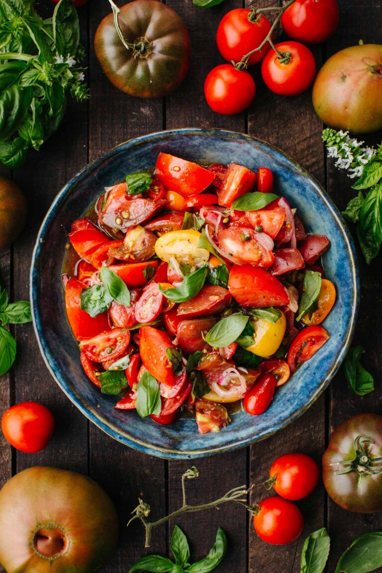 A blue ceramic bowl filled with marinated tomato salad in a dark wooden background surrounded by fresh basil and tomatoes.