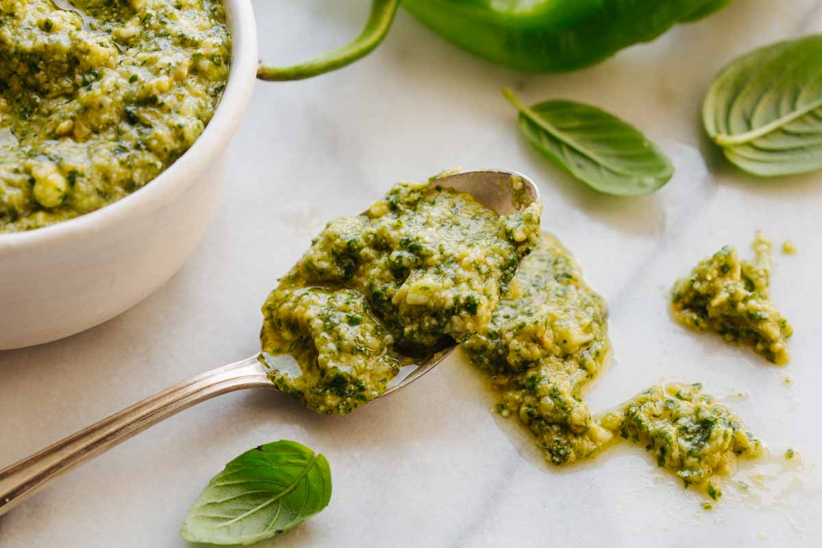Close-up shot of a spoonful of long hot pepper pesto lifted from the bowl, with fresh basil leaves and a whole long hot pepper in the background.