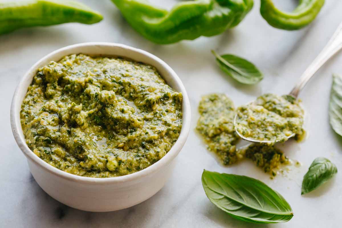 Close-up shot of a white bowl of long hot pepper pesto with a spoon, surrounded by fresh long hot peppers and basil leaves on a marble surface.
