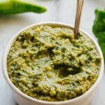 Close-up shot of long hot pepper pesto in a white bowl with a spoon standing upright in the thick, glossy pesto, basil leaves blurred in the background.