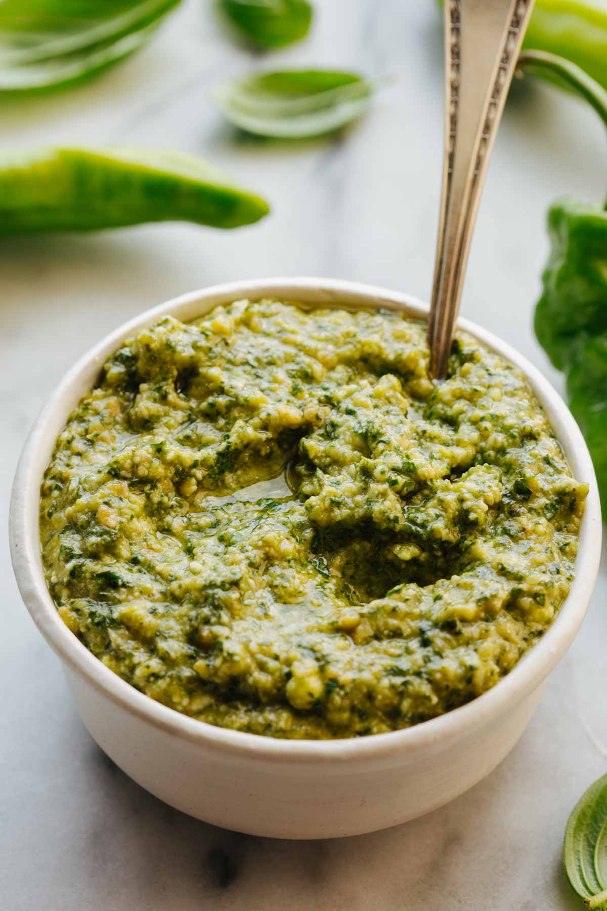 Close-up shot of long hot pepper pesto in a white bowl with a spoon standing upright in the thick, glossy pesto, basil leaves blurred in the background.