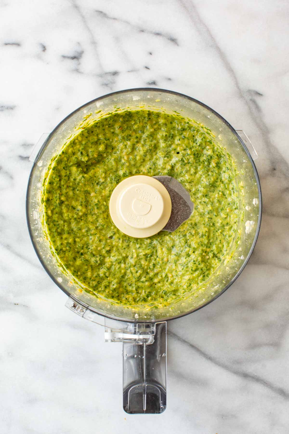 Overhead shot of long hot pepper pesto in a food processor, showing the roughly blended bright green mixture with visible flecks of pepper skin and basil before the cheese is added.