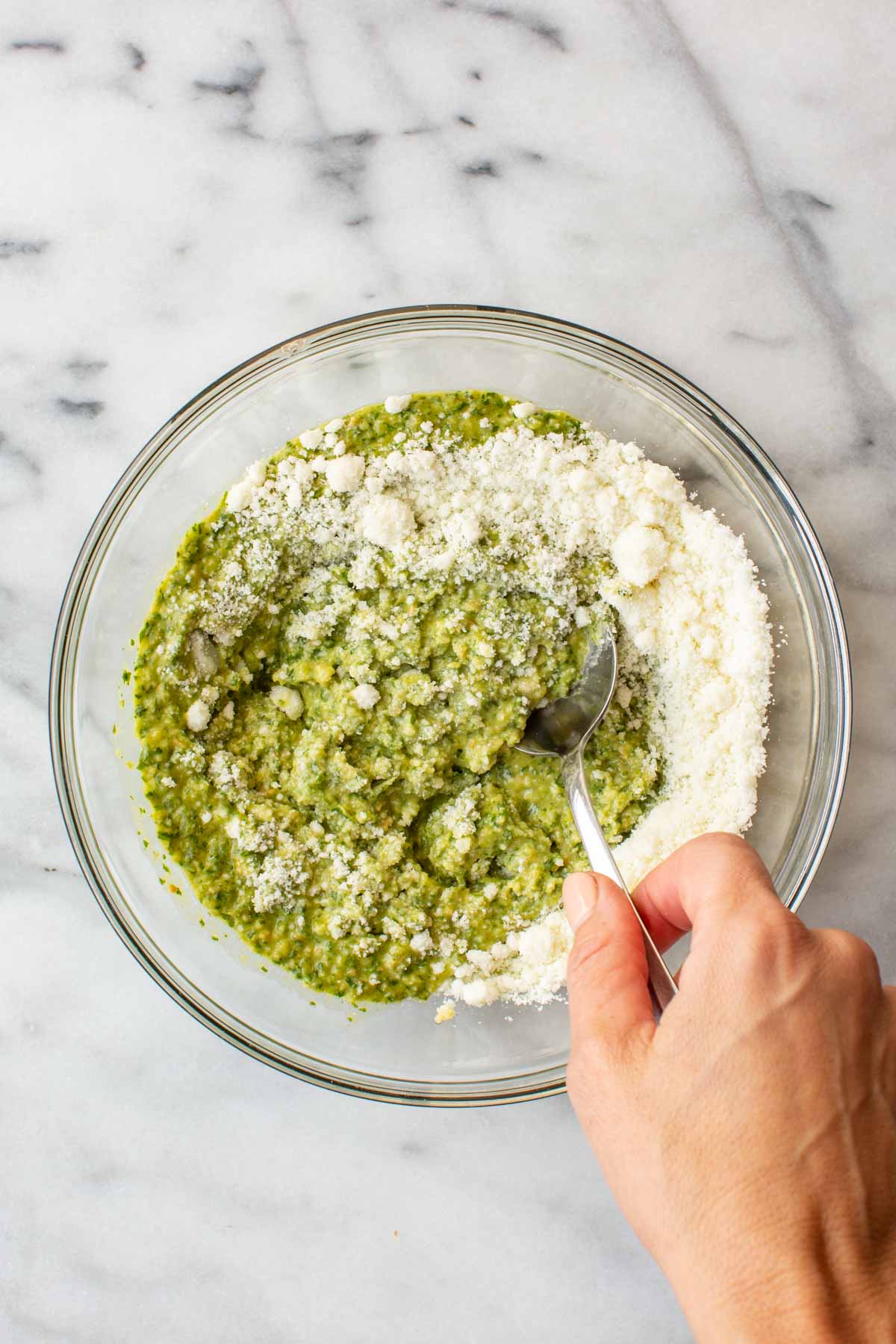 Overhead shot of a glass bowl with long hot pepper pesto being mixed with grated pecorino cheese, a spoon folding the cheese into the green pesto by hand.