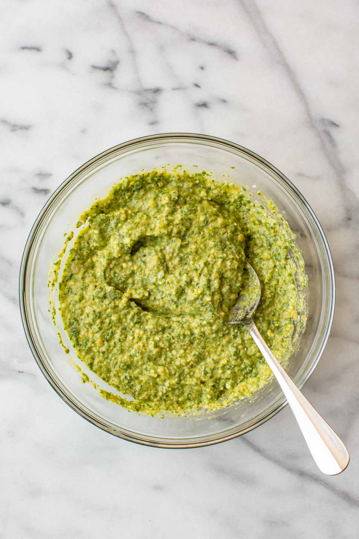 Overhead shot of the finished long hot pepper pesto in a glass bowl, fully mixed to a thick, creamy texture with a spoon resting inside.