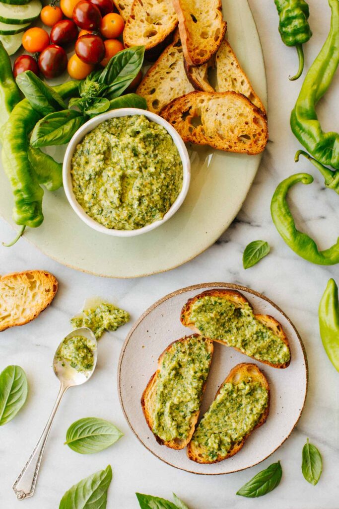 Overhead shot of a spread with a bowl of long hot pepper pesto, grilled crostini, fresh basil, cherry tomatoes, and whole long hot peppers arranged on marble, with a small plate of pesto crostini in the foreground.