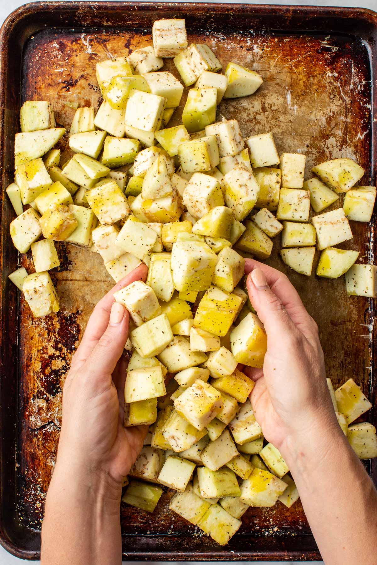 Hands tossing cubed eggplant with oregano and garlic powder on a baking sheet.