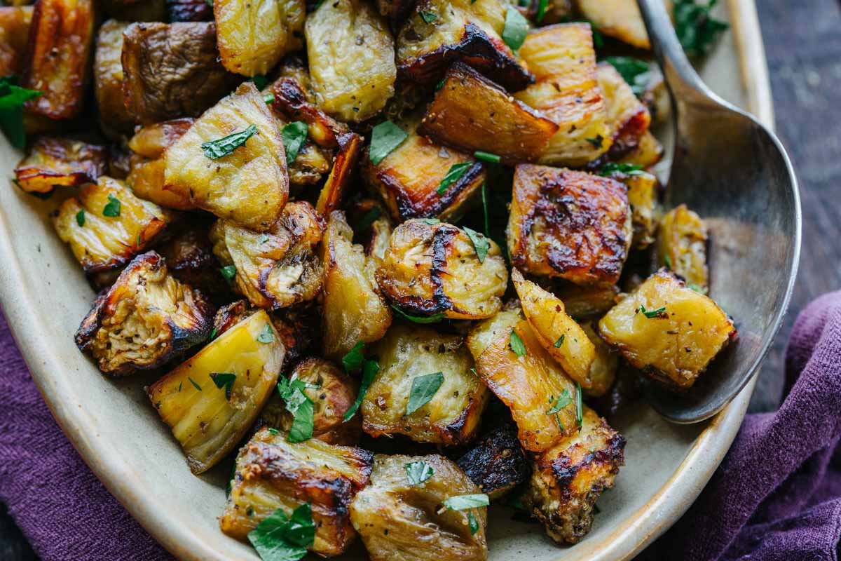Golden brown roasted eggplant pieces garnished with parsley, serving spoon resting on platter.